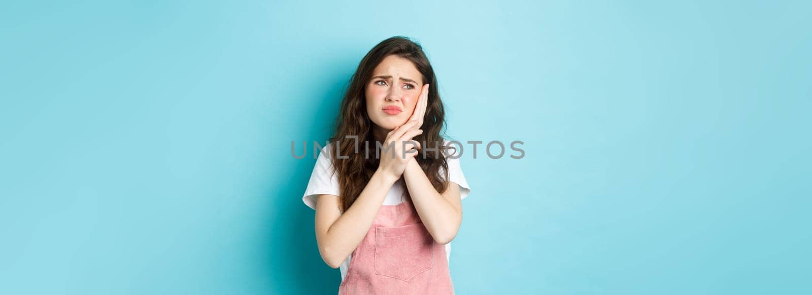 Portrait of frowning girl touching cheek, having toothache, looking at upper left corner stomatology clinic banner, suffering tooth decay, standing against blue background.