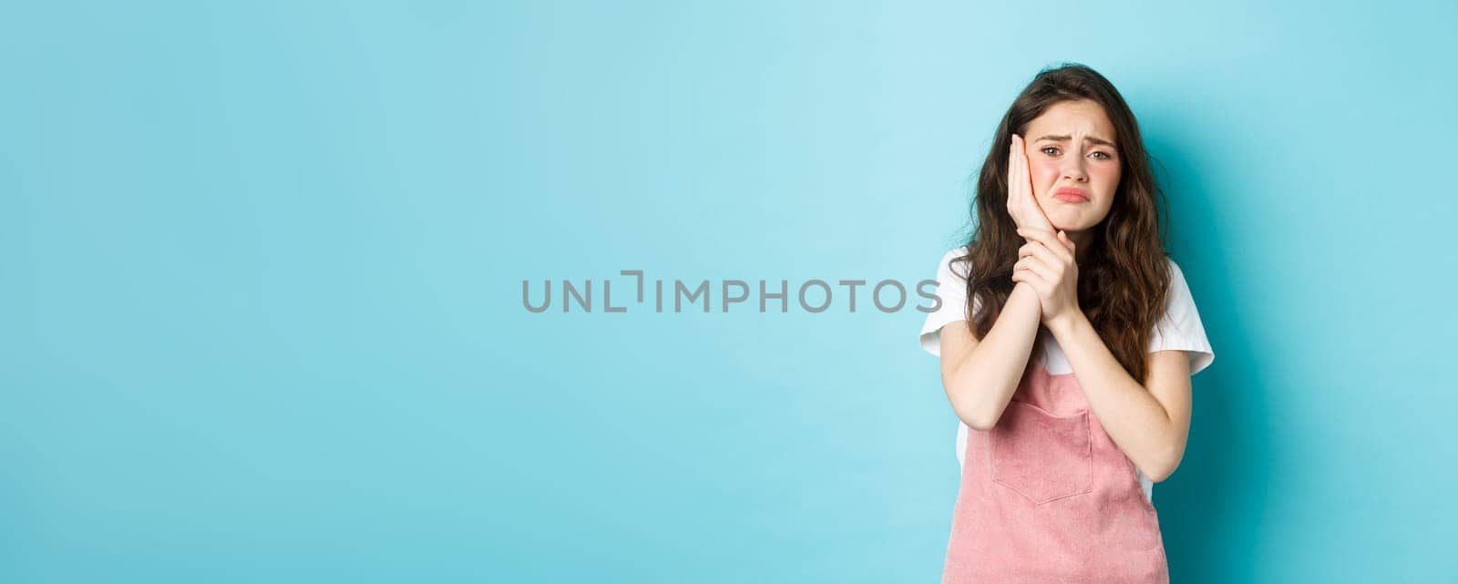 Image of frowning upset girl touching swallen cheek, having toothache, complaining on tooth decay, need to see dentist, standing against blue background.