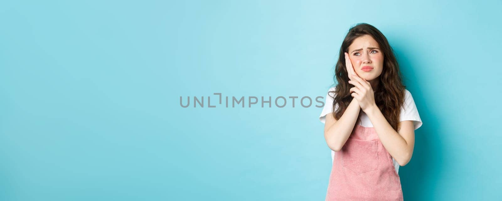 Portrait of frowning girl touching cheek, having toothache, looking at upper left corner stomatology clinic banner, suffering tooth decay, standing against blue background.