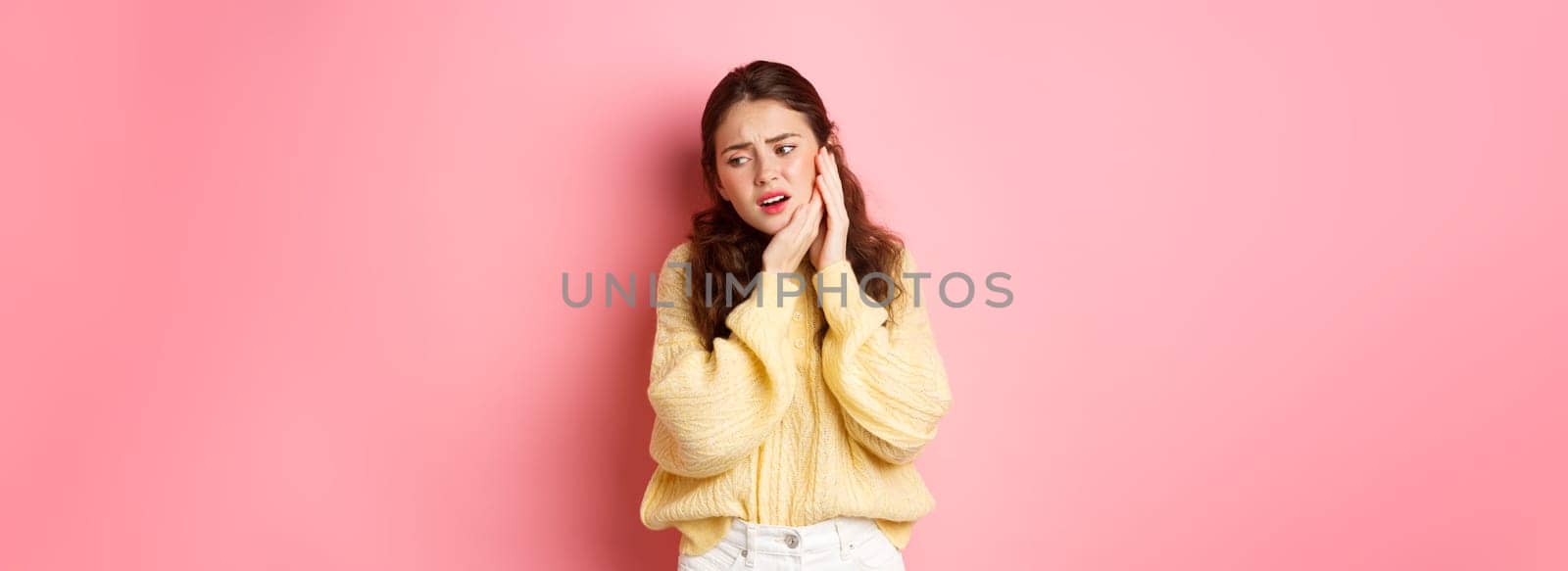 Young woman having toothache, touching swallen cheek and grimacing from pain, need to see dentist, stomatology appointment, standing against pink background.