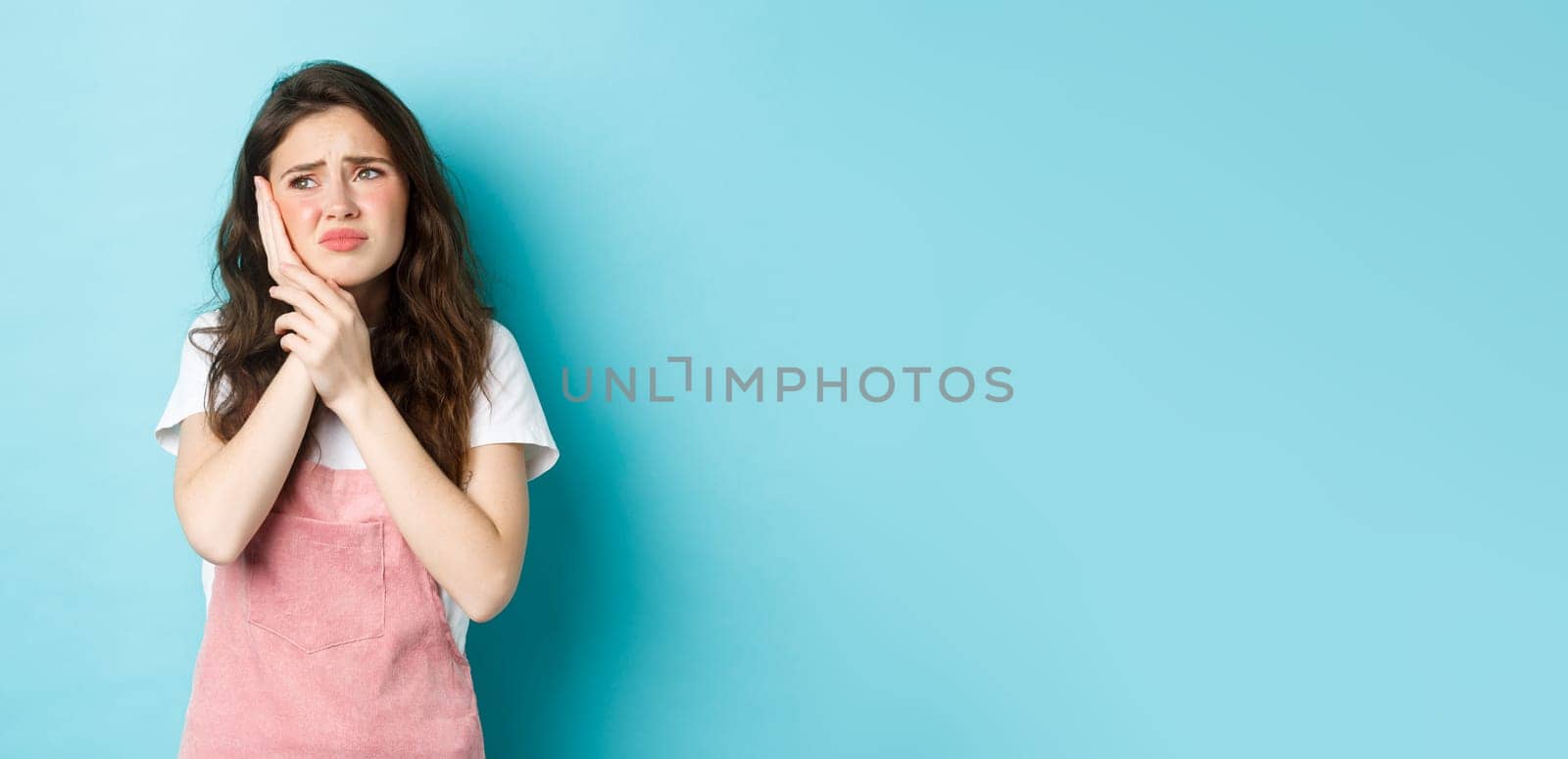 Portrait of frowning girl touching cheek, having toothache, looking at upper left corner stomatology clinic banner, suffering tooth decay, standing against blue background.