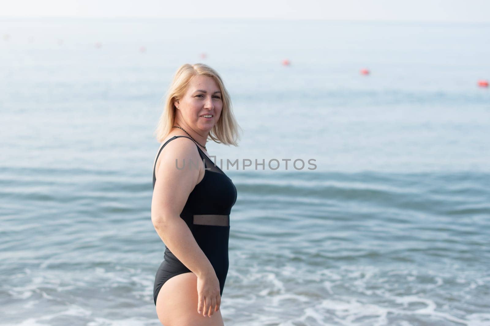 Young blonde woman with beautiful flowing hair in fashionable closed black swimsuit posing against the background of the sea on the seashore during summer vacation, copy space