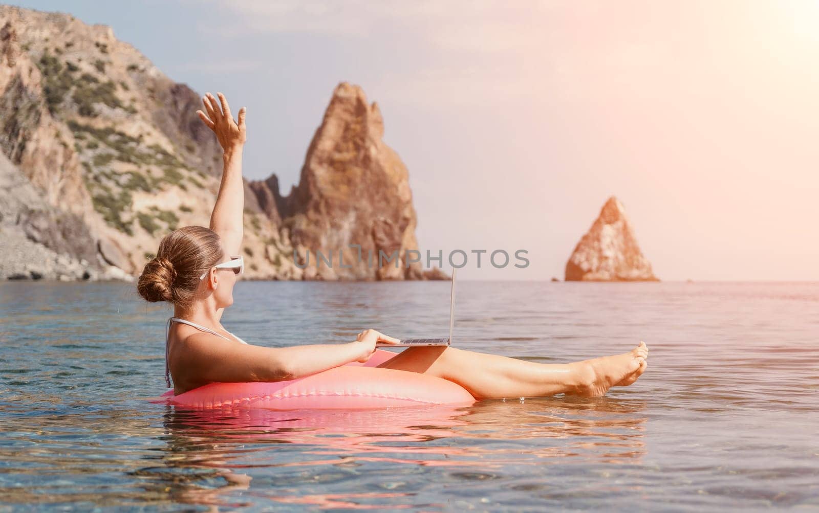 Woman freelancer works on laptop swimming in sea on pink inflatable ring. Pretty lady typing on computer while floating in the sea on inflatable donut at sunset. Freelance, remote work on vacation