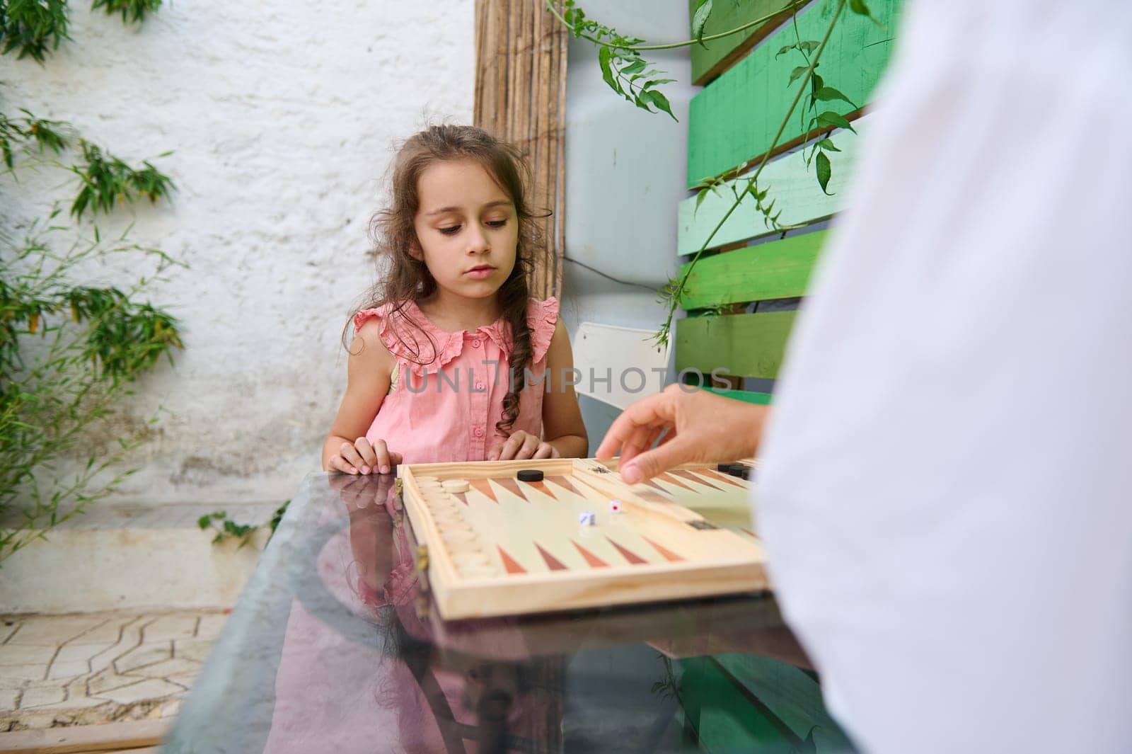 Young girl in a pink dress concentrating on learning to play backgammon with family in an outdoor summer setting.