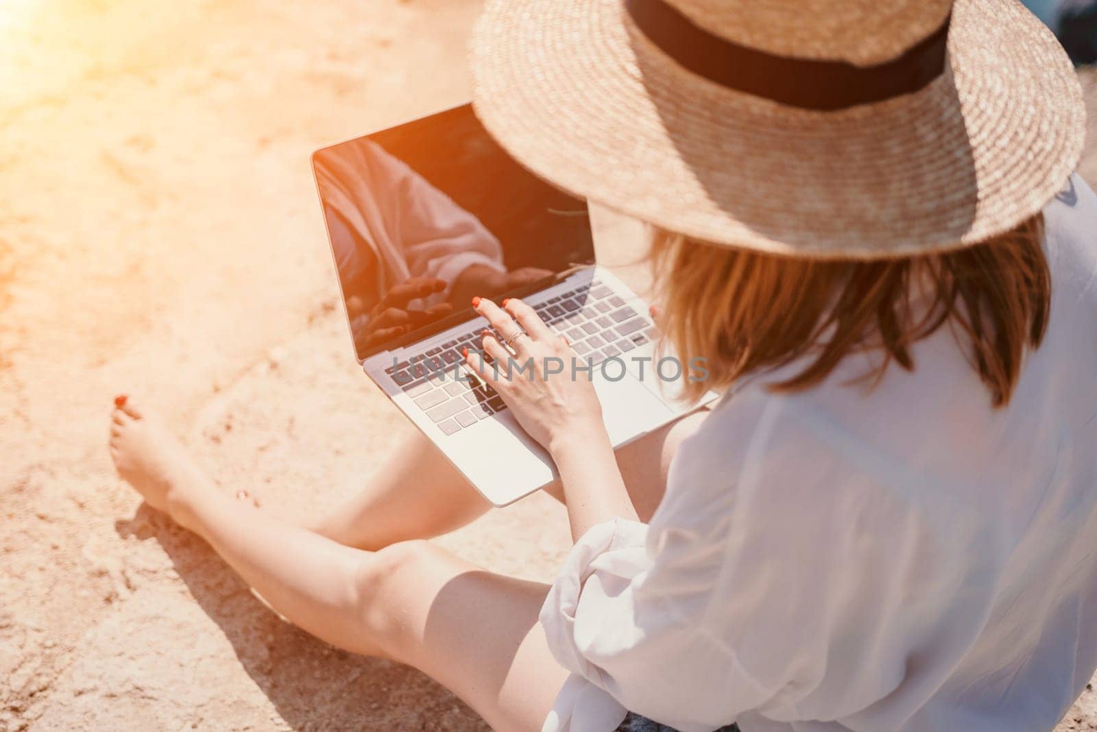 Successful business woman in yellow hat working on laptop by the sea. Pretty lady typing on computer at summer day outdoors. Freelance, travel and holidays concept.