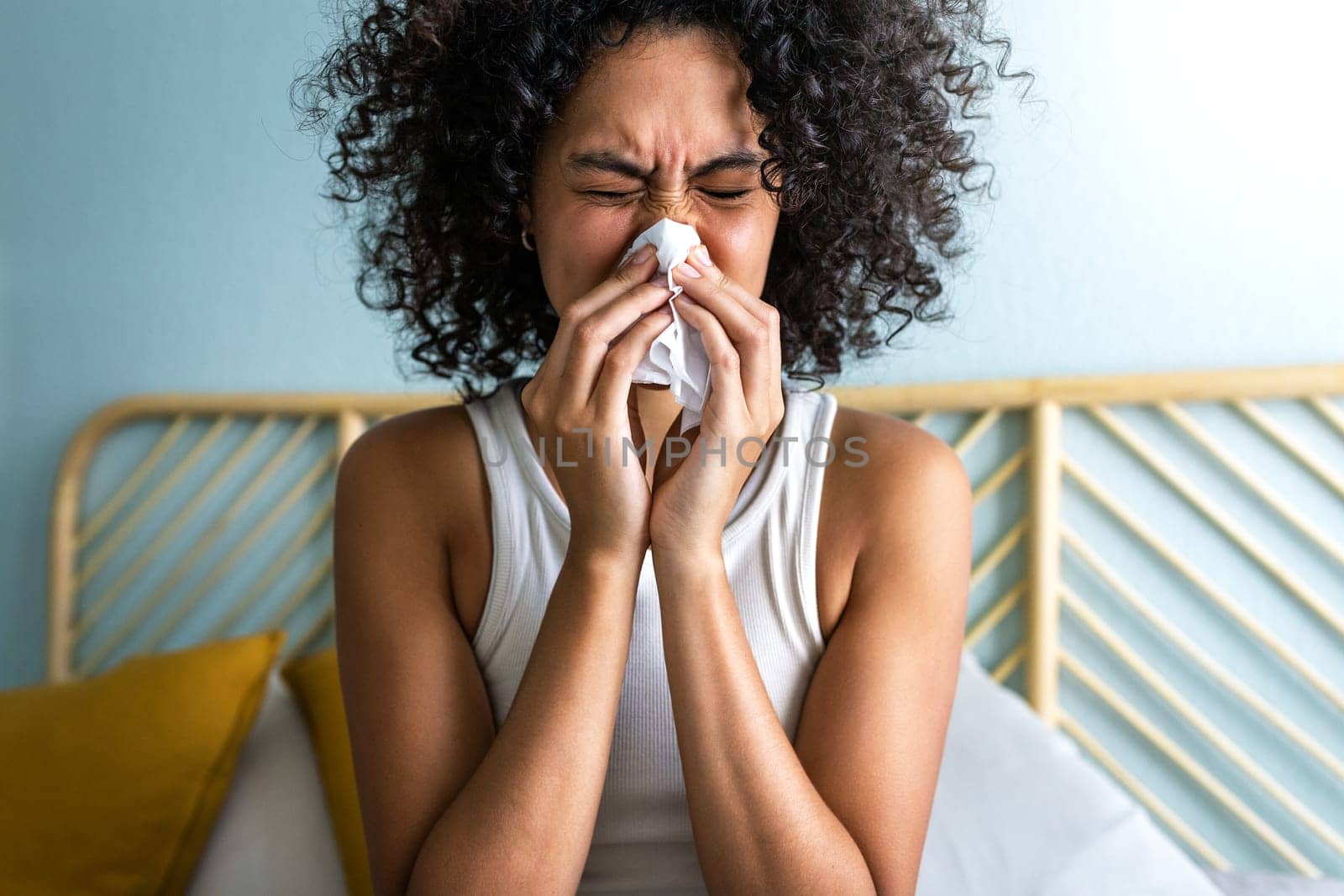 Young multiracial woman blowing nose at home sitting on bed. Sickness concept.