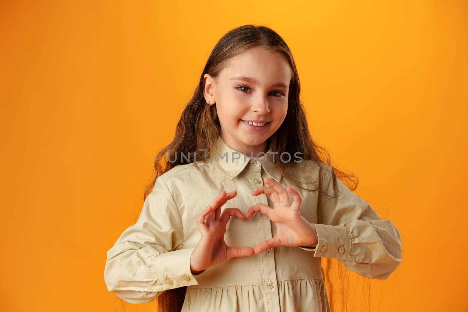 Happy little girl making heart shape by hands on yellow background in studio