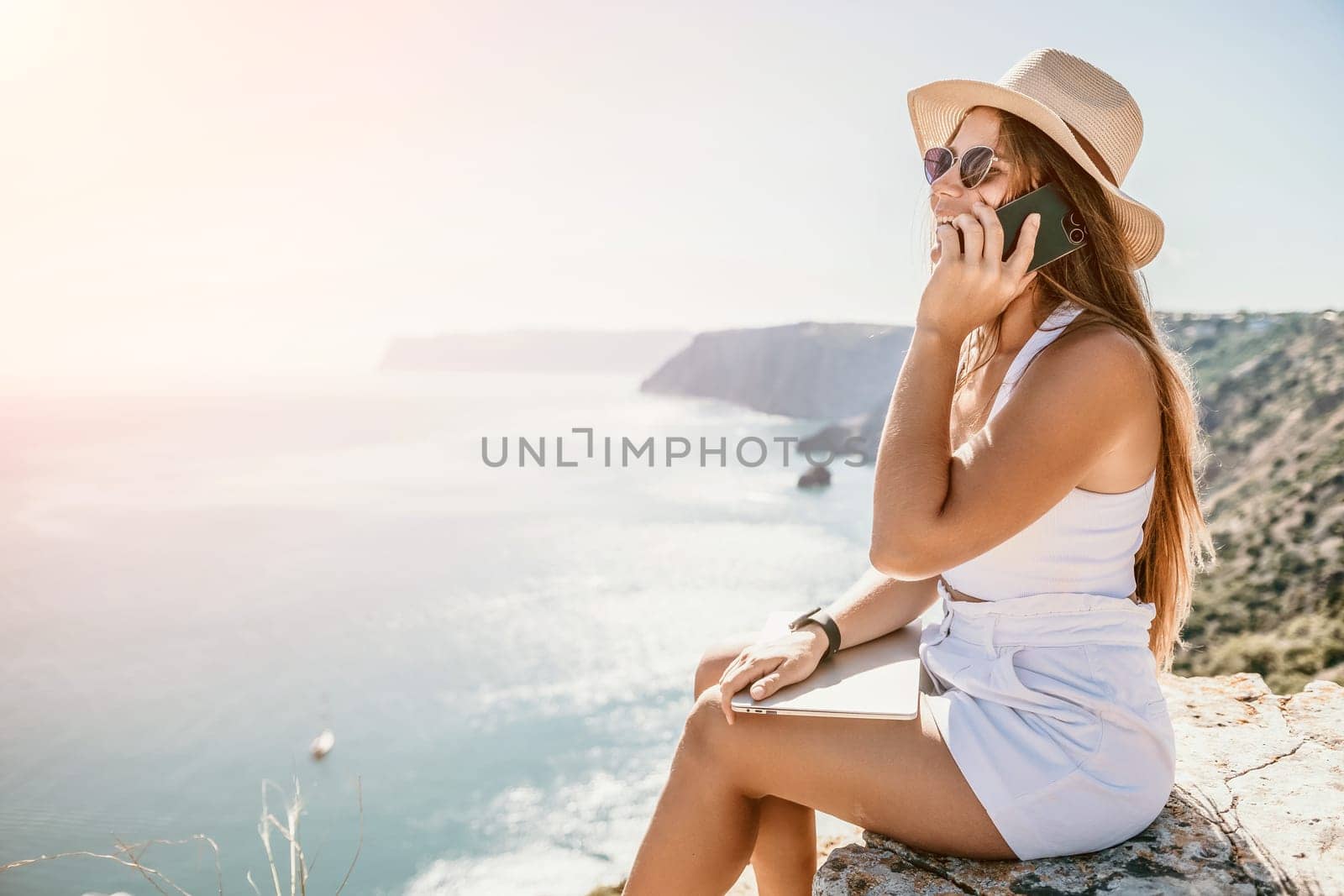 Successful business woman in yellow hat working on laptop by the sea. Pretty lady typing on computer at summer day outdoors. Freelance, travel and holidays concept.