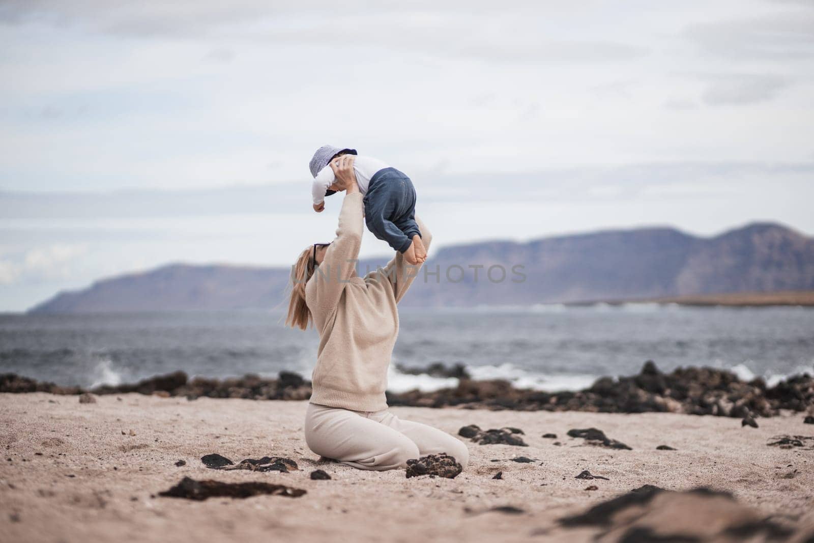 Mother enjoying winter vacations holding, playing and lifting his infant baby boy son high in the air on sandy beach on Lanzarote island, Spain. Family travel and vacations concept by kasto