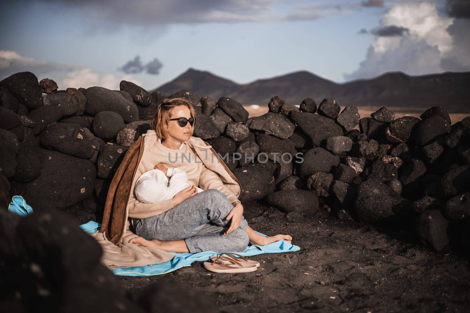 Mother breast feeding his infant baby boy son on black sandy volcanic beach of Janubio on Lanzarote island, Spain, enjoing dramatic volcanic landscape on windy overcast day. Travel with kids concept by kasto