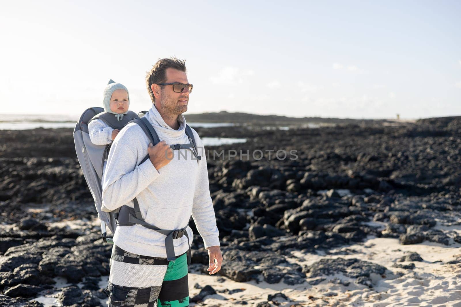 Young father carrying his infant baby boy son in backpack on black rock volcanic beach on Lanzarote island, Spain. Family travel and winter vacation concept. by kasto