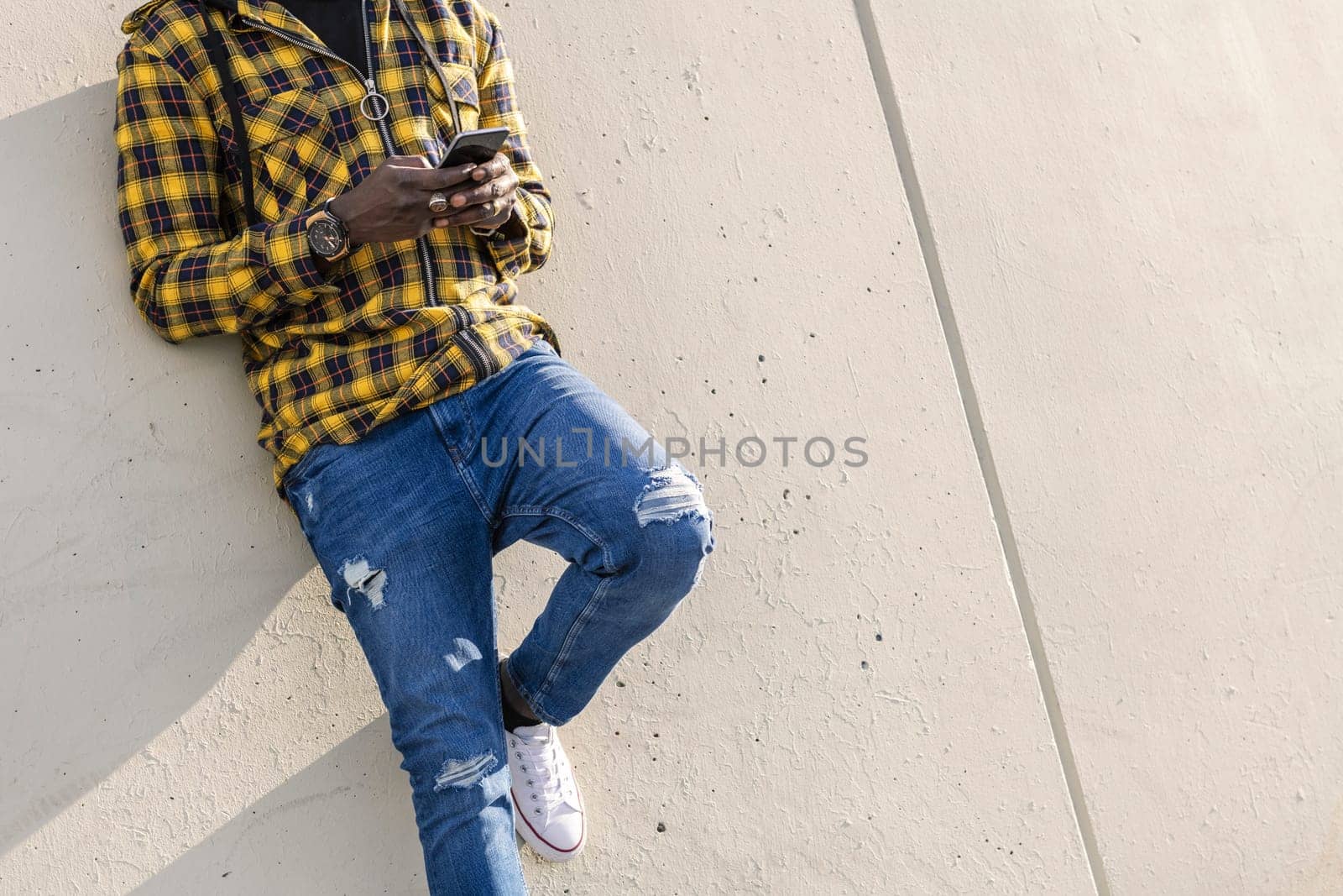 unrecognizable young african man writing a message on his phone leaning on a modern concrete structure, concept of lifestyle and technology