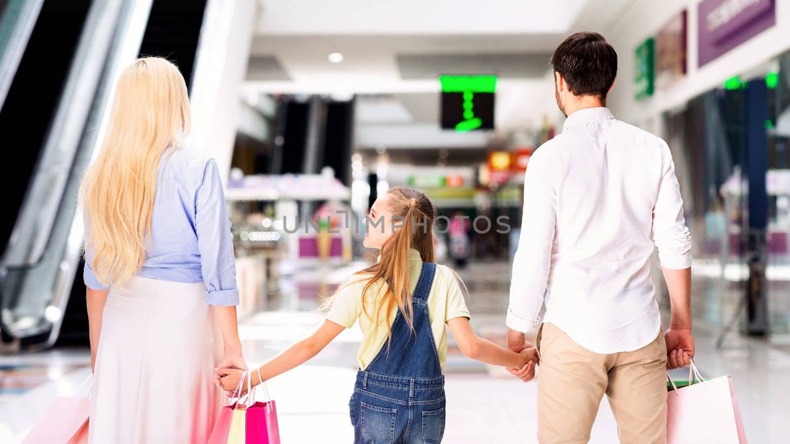 A family of three walks through a shopping mall. They are holding hands, and the parents each carry a shopping bag. The young girl is looking back at something behind them