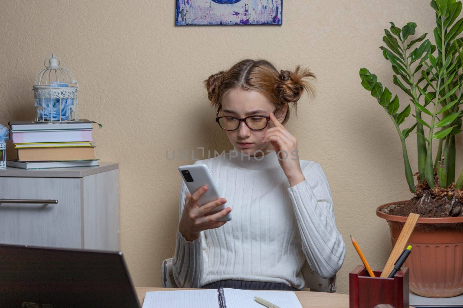 beautiful girl in glasses looks thoughtfully at the smartphone screen while sitting at her desk. soft focus