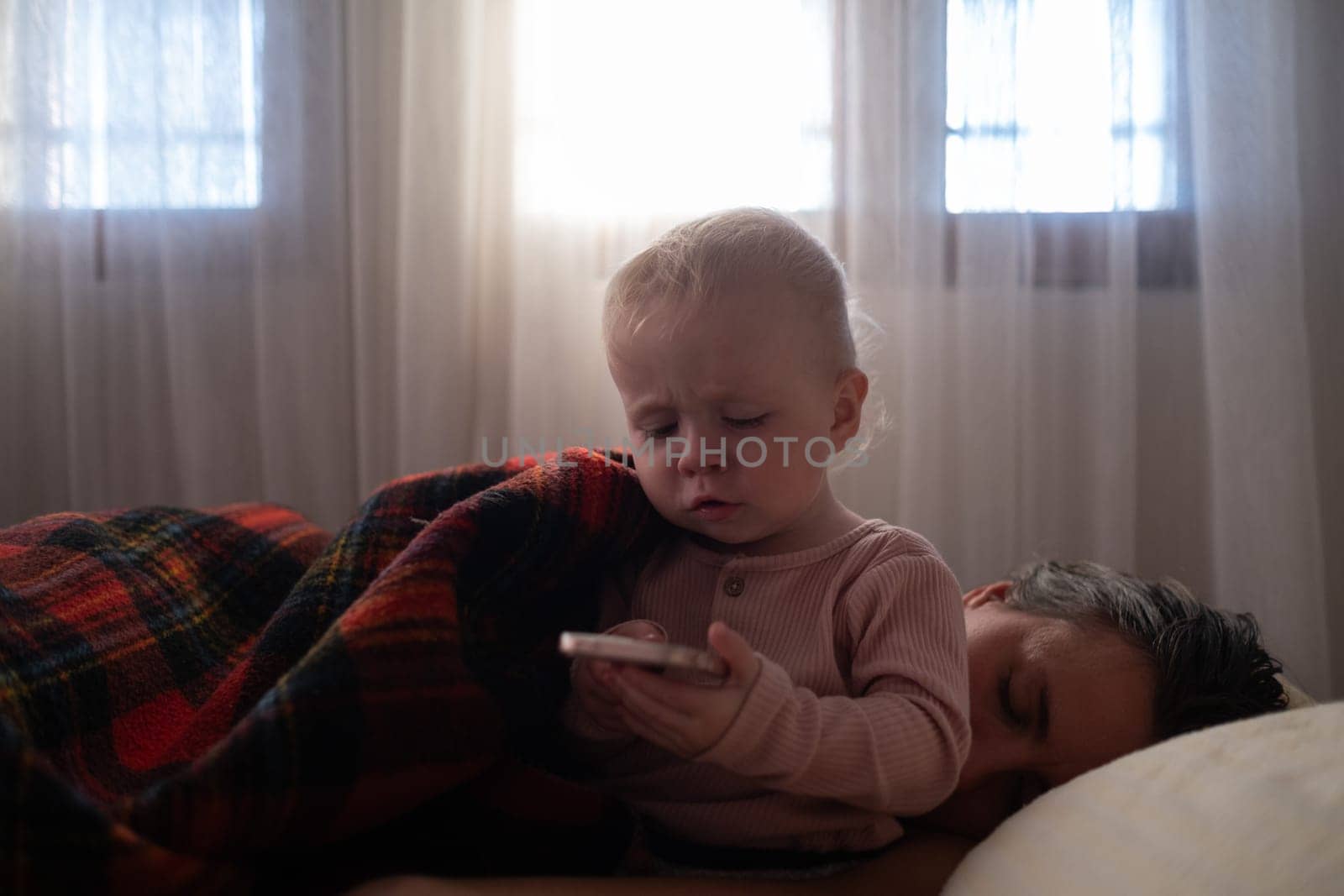 A 2 year old girl plays games on phone while lying in bed. Gadgets in bed before bedtime in a child. Mother is tired and sleeping