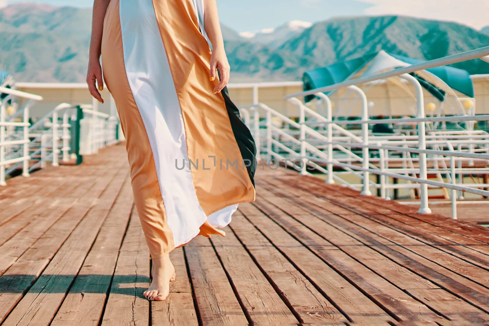 Legs of a woman in a dress walking along the pier on the beach by the sea in a hotel while relaxing against the backdrop of high mountains