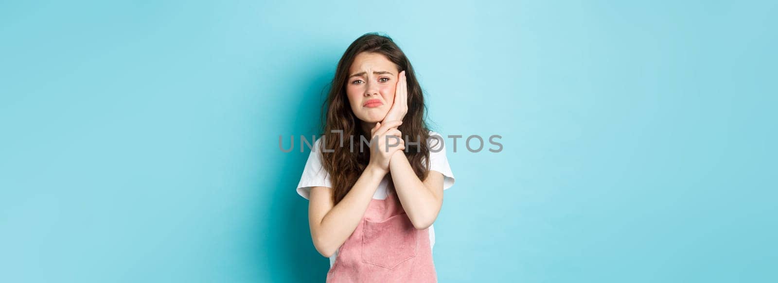 Image of frowning upset girl touching swallen cheek, having toothache, complaining on tooth decay, need to see dentist, standing against blue background.