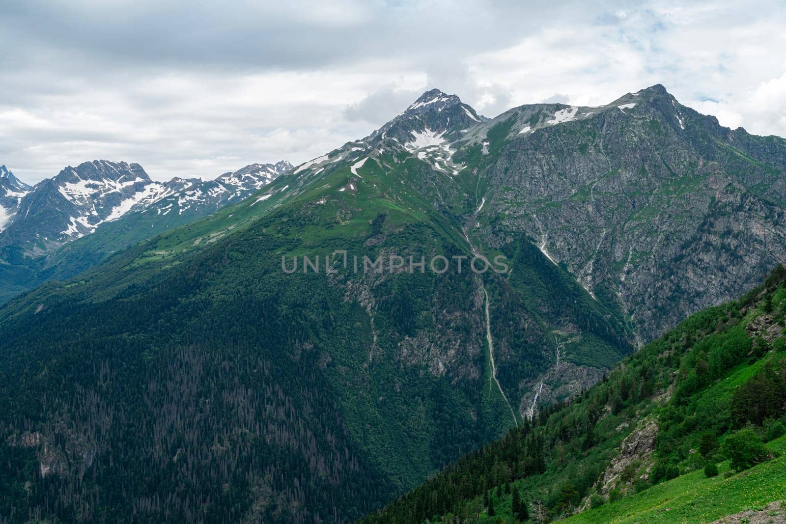 A drone flies over a mountain valley in the Caucasus Mountains. The valley is filled with green trees and a winding river. In the distance, a snow-capped peak rises above the valley.