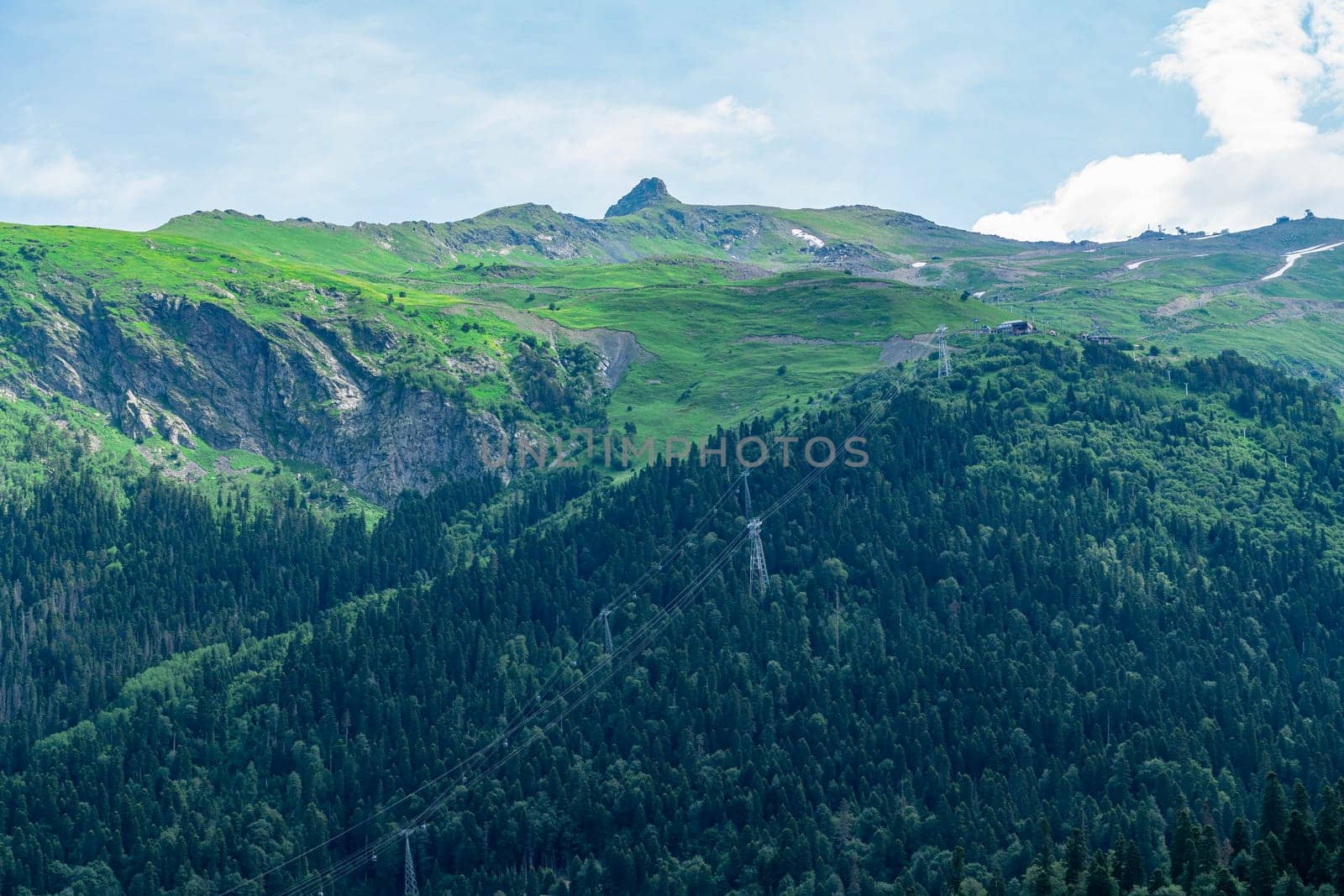 A drone flies over a mountain valley in the Caucasus Mountains. The valley is filled with green trees and a winding river. In the distance, a snow-capped peak rises above the valley.