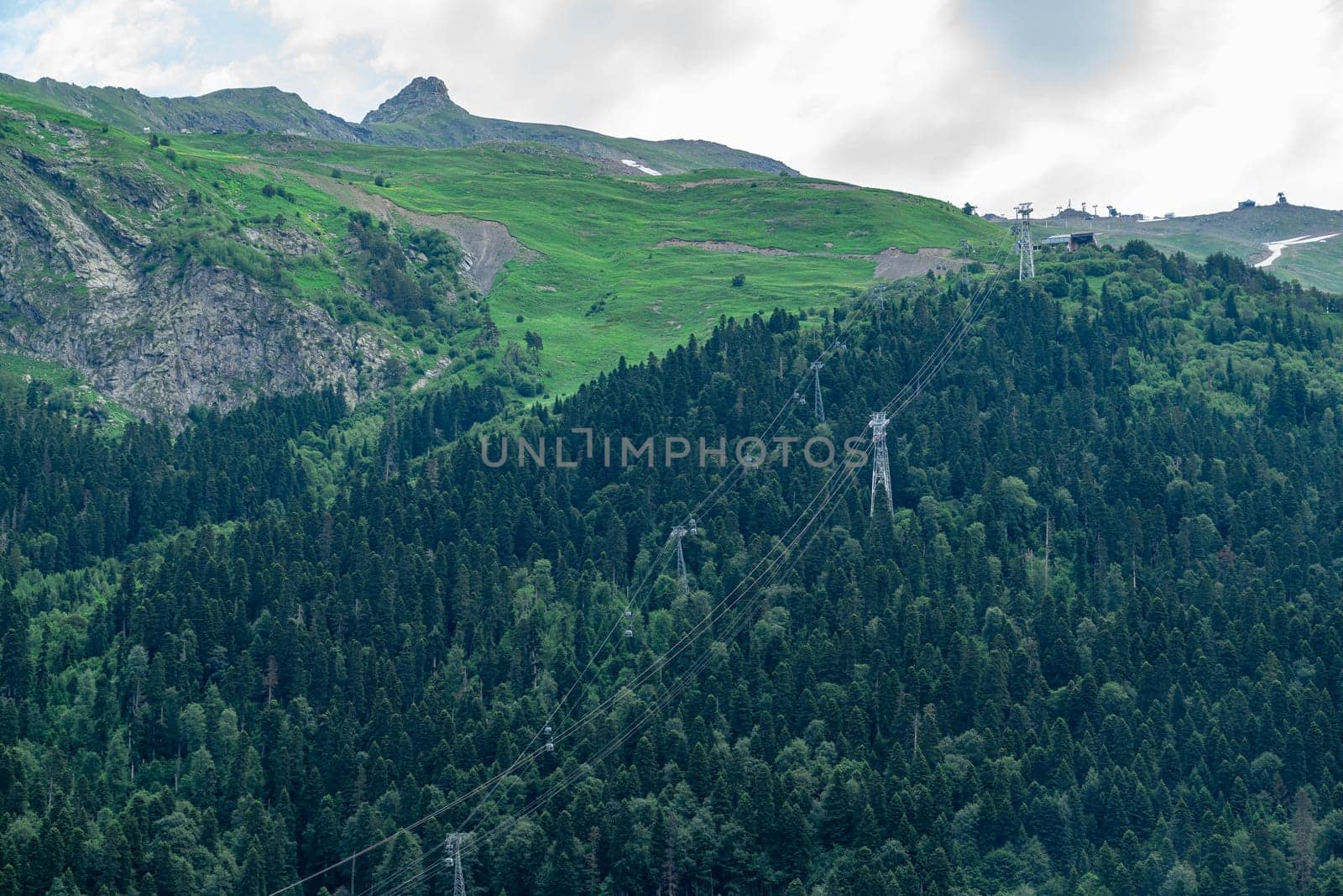 A drone flies over a mountain valley in the Caucasus Mountains. The valley is filled with green trees and a winding river. In the distance, a snow-capped peak rises above the valley.