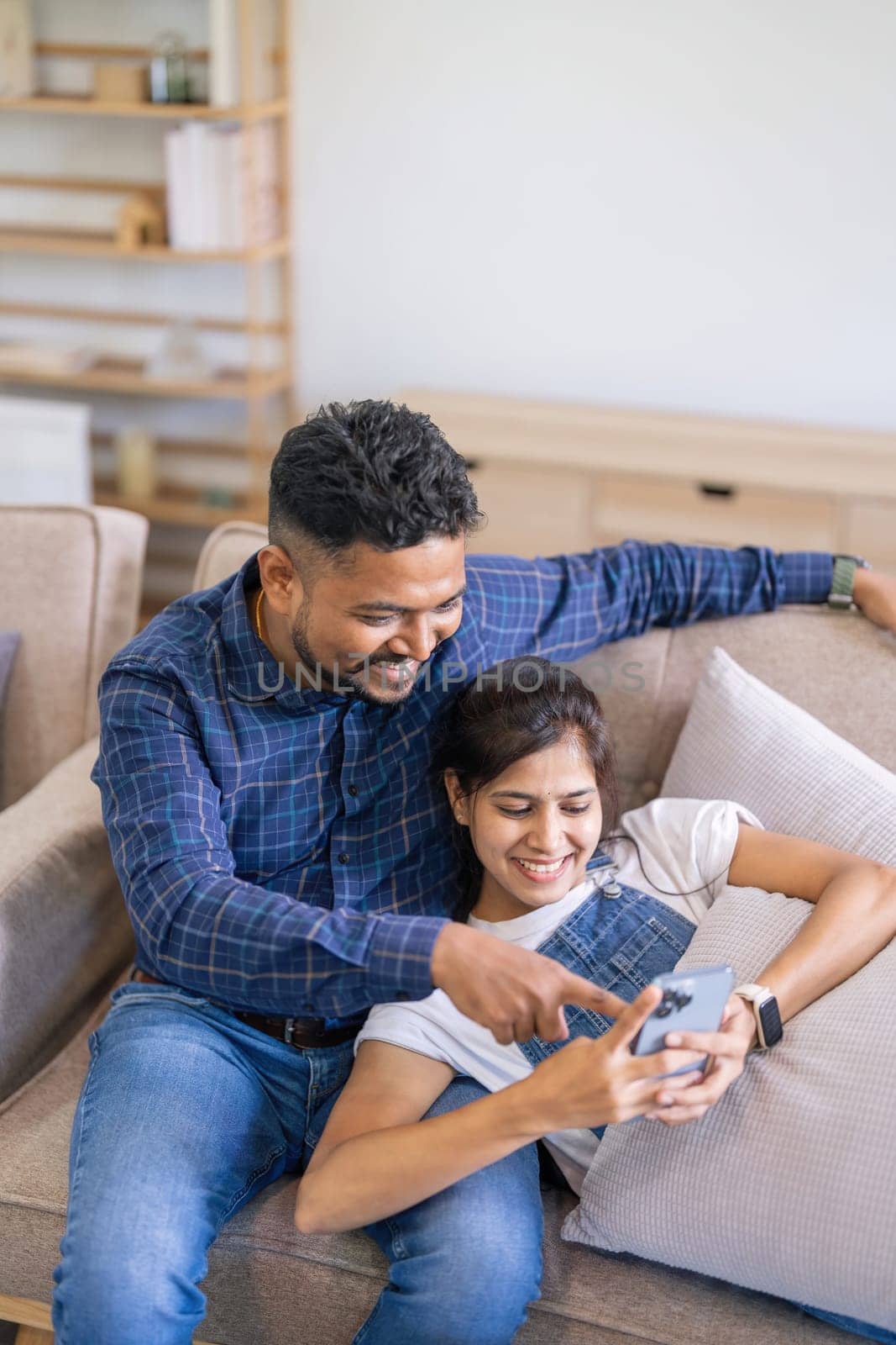 Happy young indian family couple using smart phone looking at cellphone sit on sofa together relax at home check social media apps or shopping in mobile application.