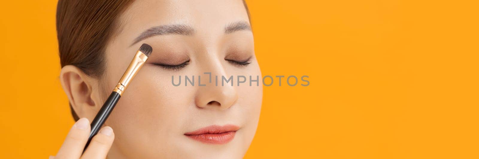 Yellow banner studio photo of a young woman using make up brush and eye shadows.