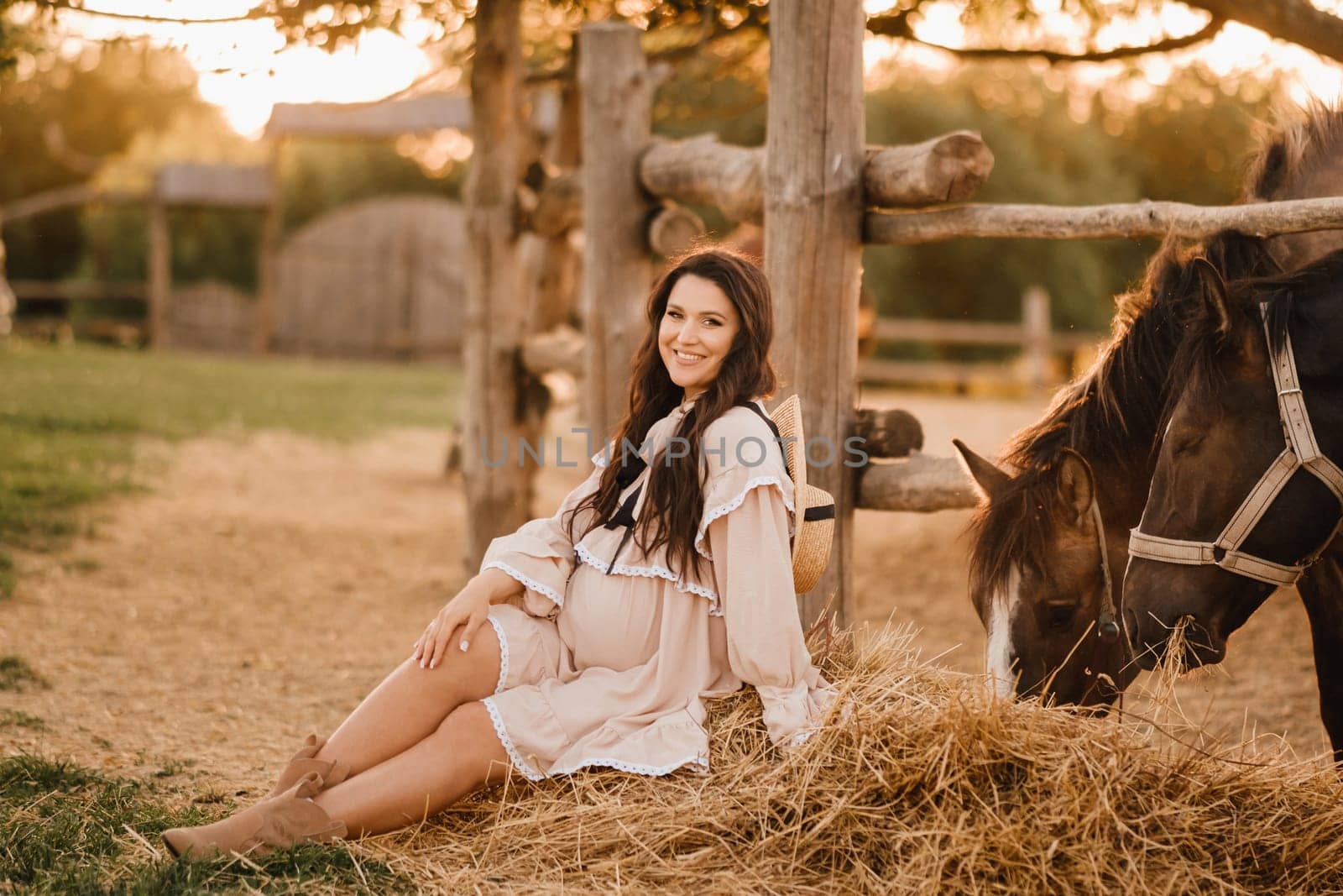 a pregnant woman in a dress in the countryside is sitting on the hay near the horses by Lobachad