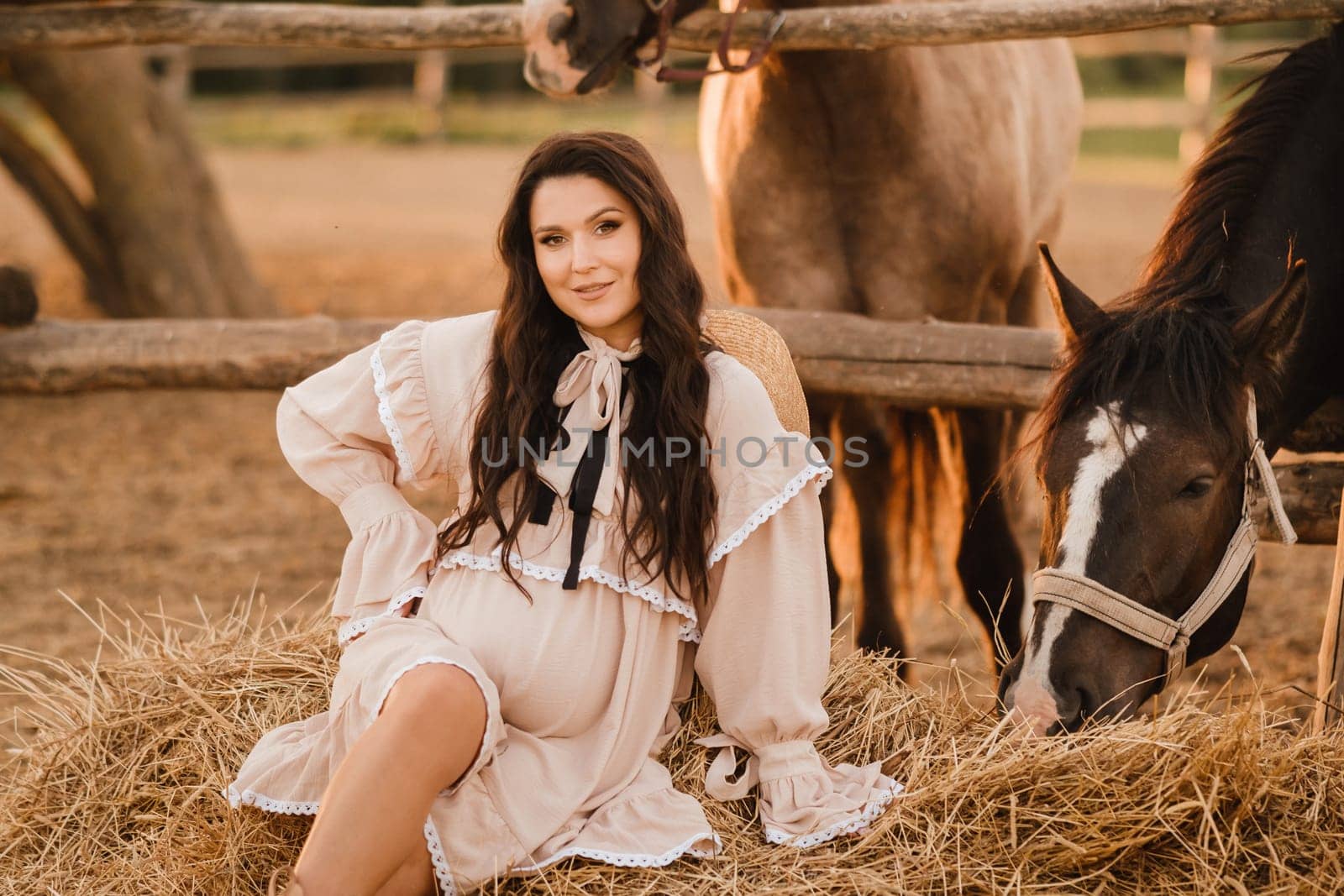 a pregnant woman in a dress in the countryside is sitting on the hay near the horses.