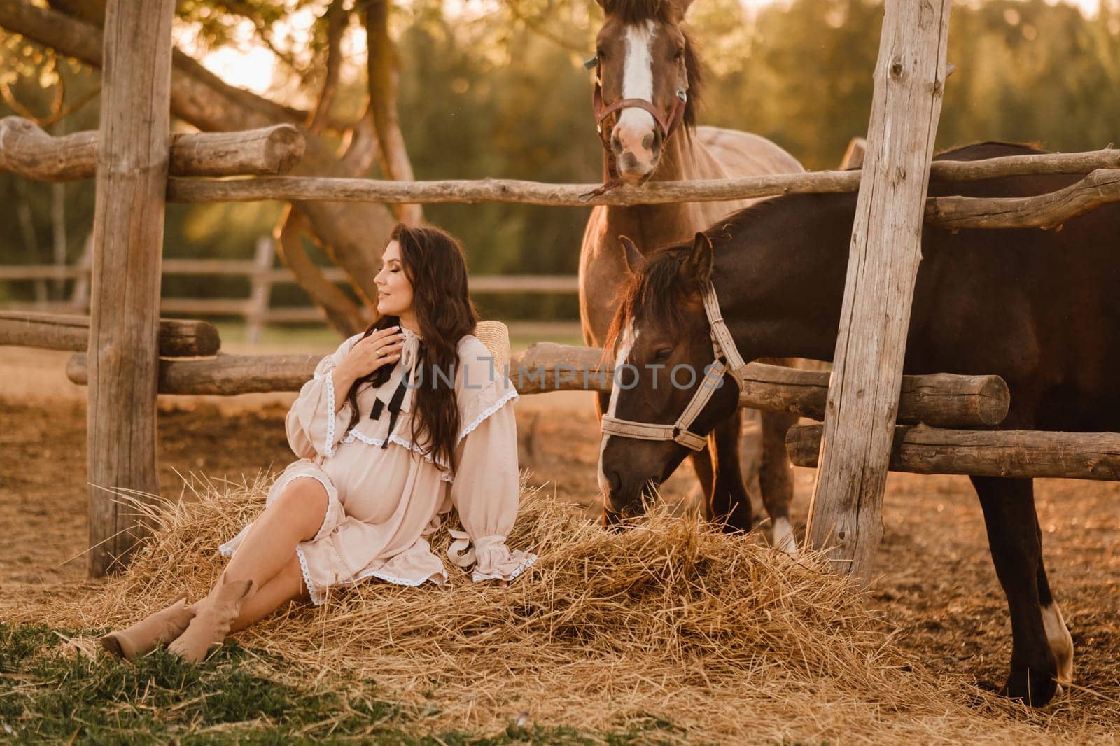 a pregnant woman in a dress in the countryside is sitting on the hay near the horses.