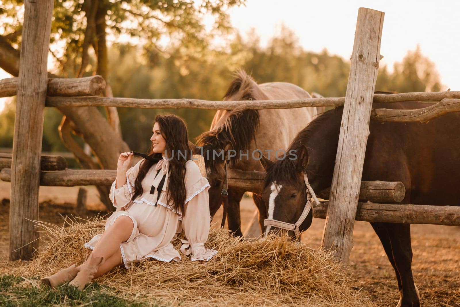 a pregnant woman in a dress in the countryside is sitting on the hay near the horses.