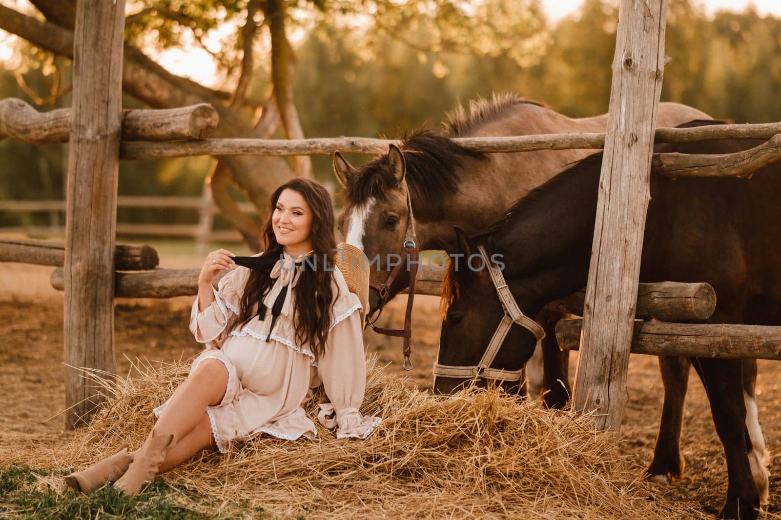 a pregnant woman in a dress in the countryside is sitting on the hay near the horses by Lobachad