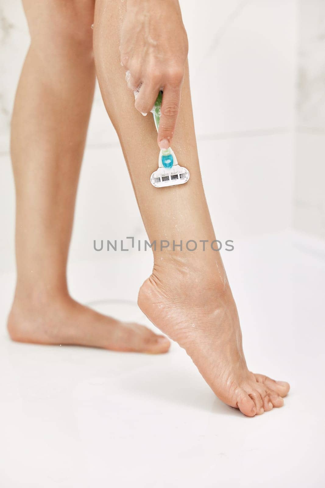 Young woman shaving her legs with razor in white shower, close-up