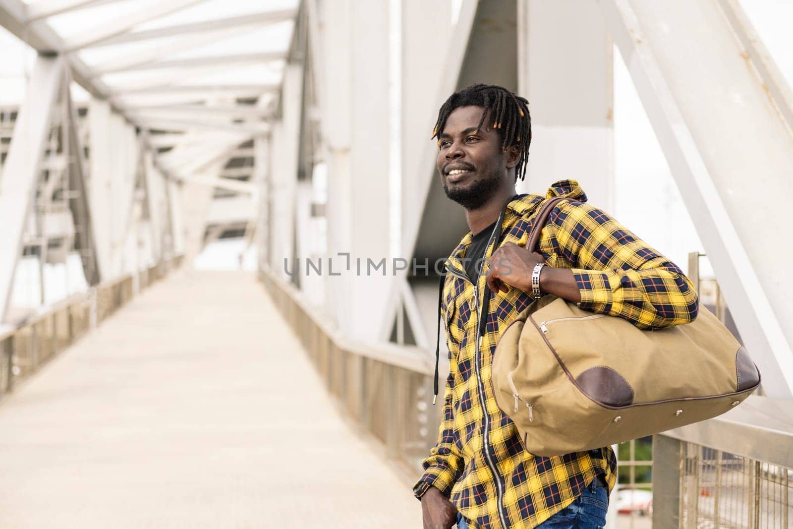 smiling young black man in front of a modern metal structure to access to the airport with a large travel bag, lifestyle and travel concept