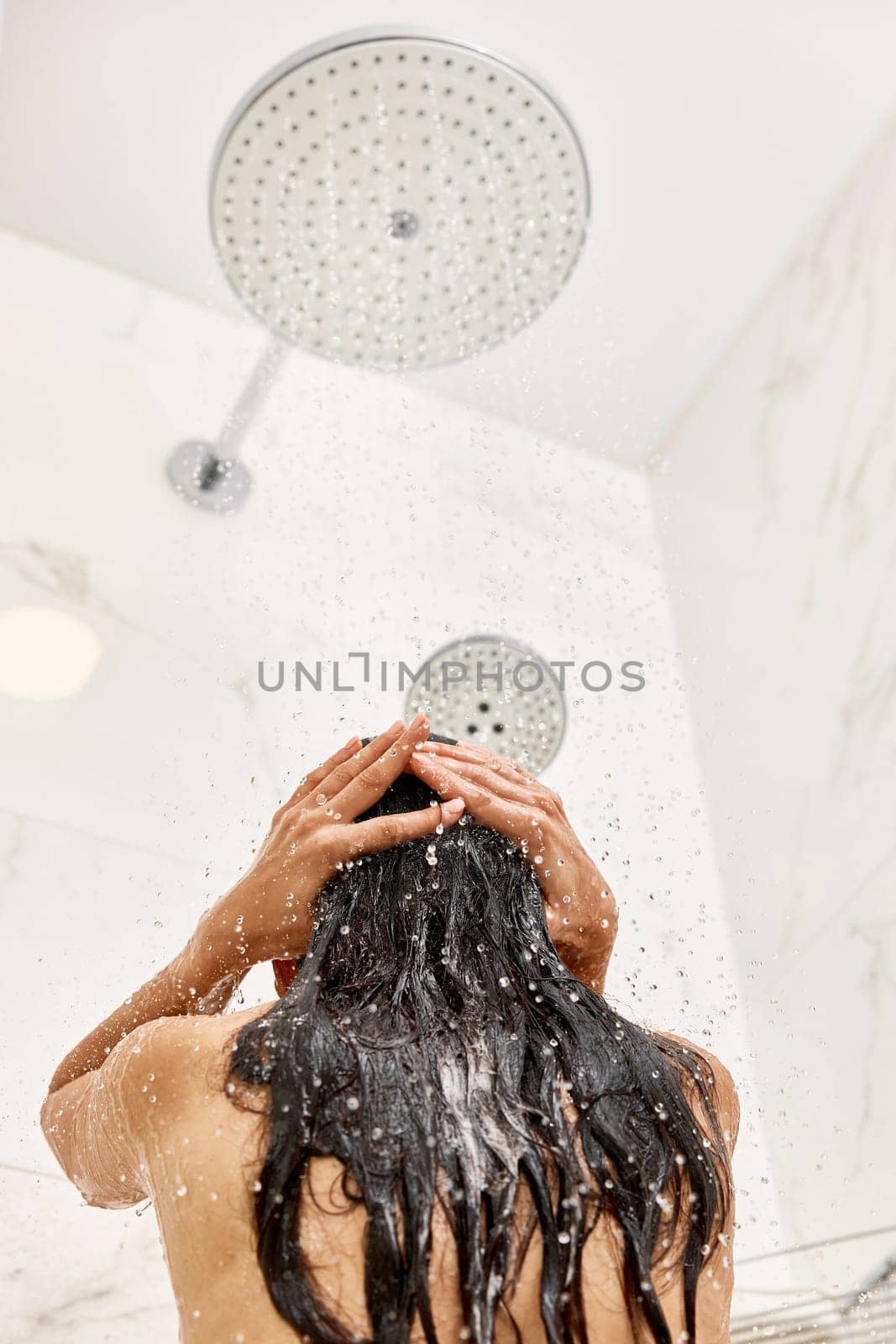 woman taking refreshing shower in marble bathroom. girl washes shampoo out of hair, back view