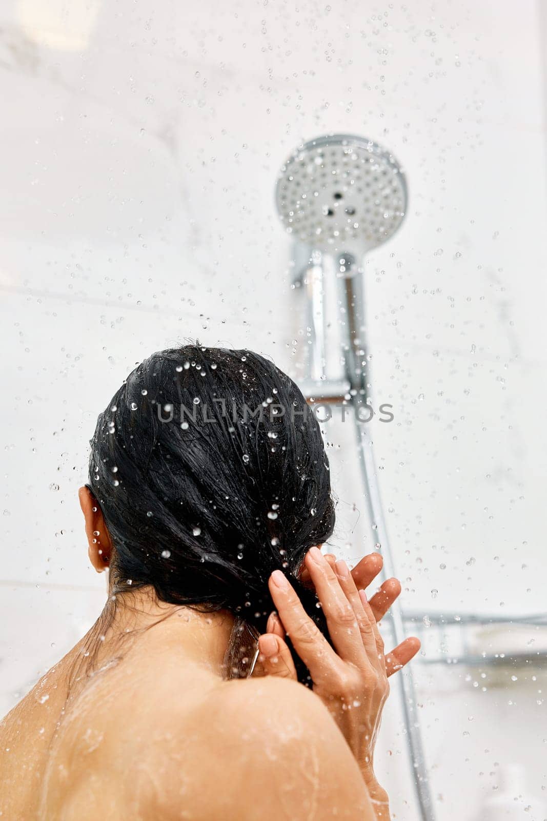 Young woman taking refreshing shower in marble bathroom. Girl washing her hair, back view