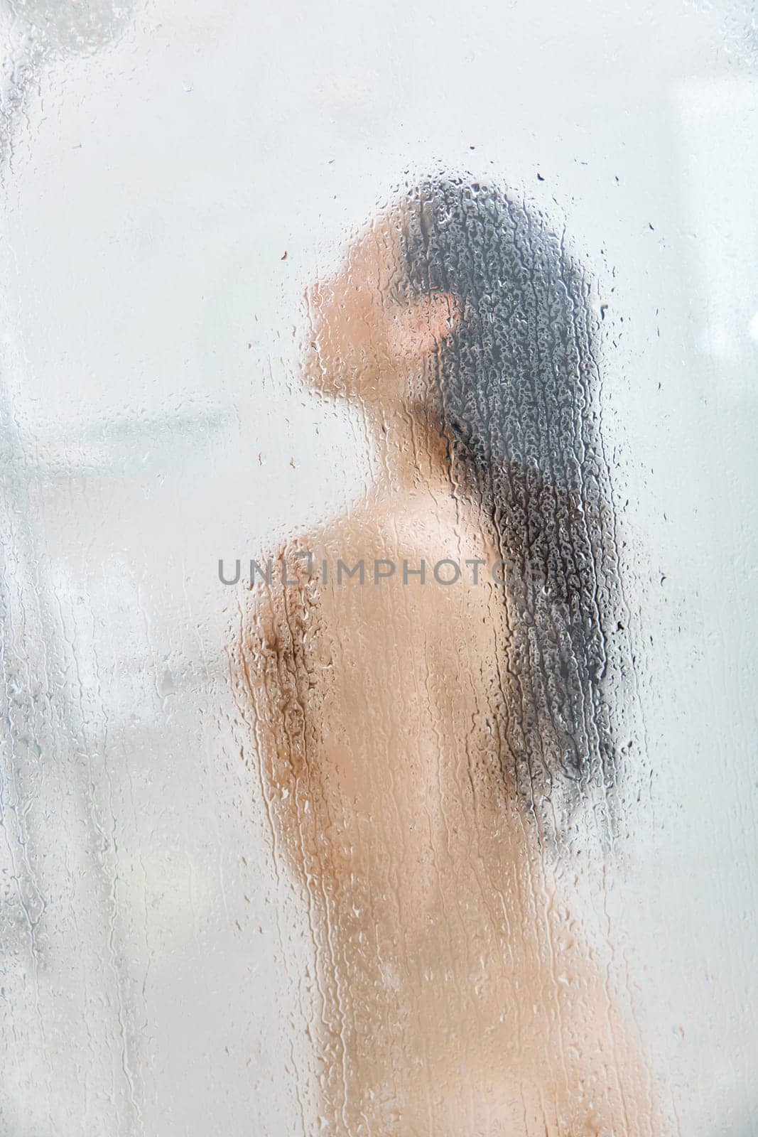 Unfocused woman washing long hair in shower behind glass with drops. Focus on drops