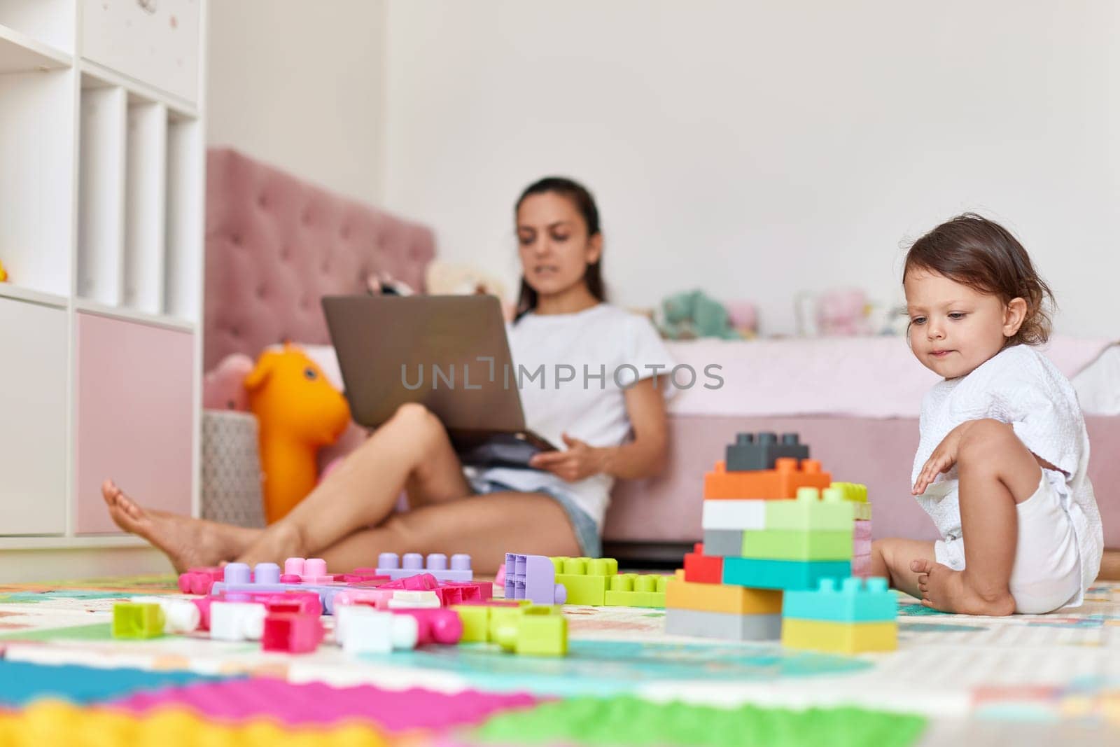 cute little baby girl playing with toys blocks and mother working on laptop from home