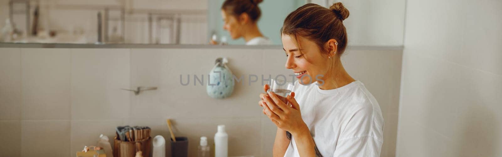 Young beautiful woman drinking glass of water in the bathroom at home. Beauty treatment