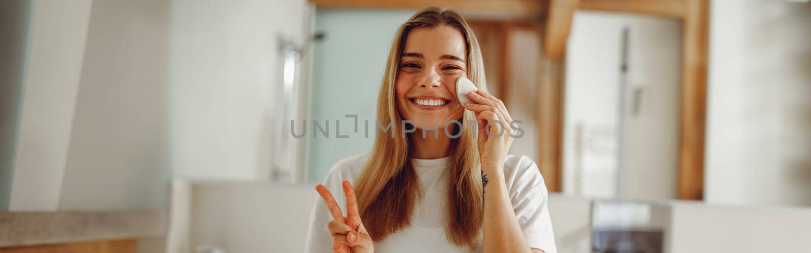 Young woman removing makeup with cotton pad standing in bathroom. Home beauty routine
