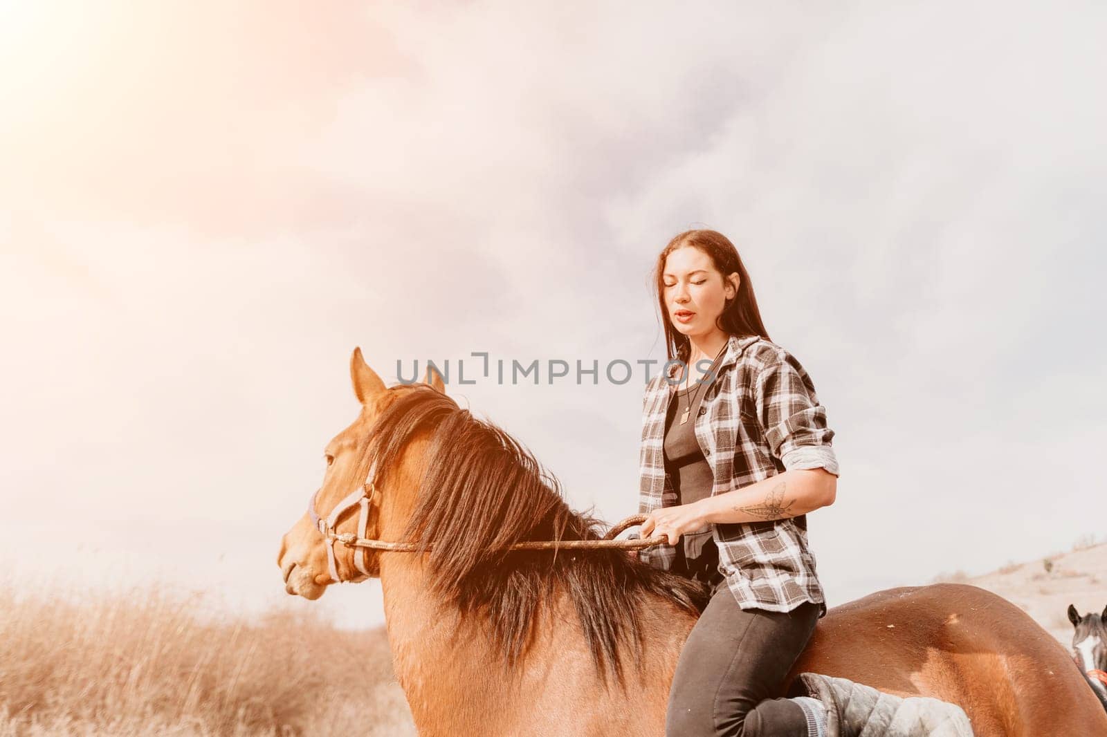 A woman in a plaid shirt rides a brown horse through a grassy field, with the sky above lit by the warm light of the sun.