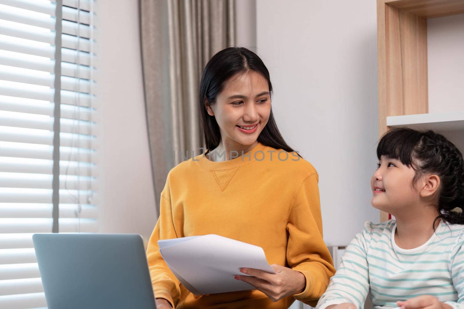 A mother and daughter share a joyful moment at home, working on a laptop and holding documents, showcasing family bonding and togetherness.