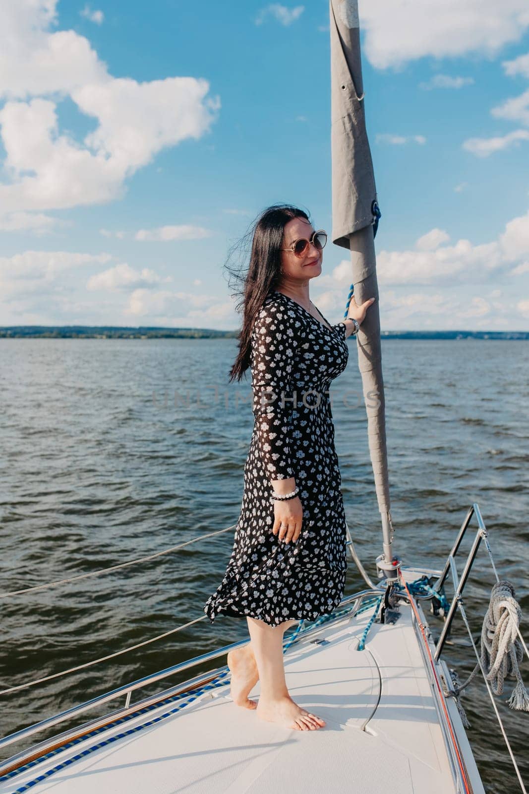 A woman in a patterned dress and sunglasses stands on a sailing boat under a clear sky