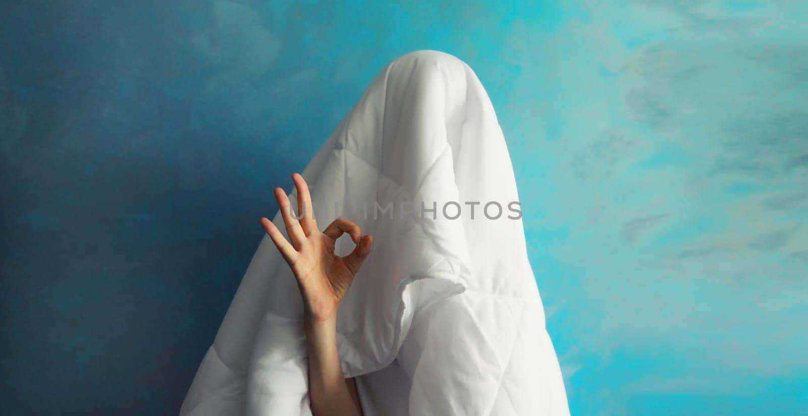 Cheerful lazy woman waking up after sleeping covered head with white blanket showing ok gesture her fingers on blue background