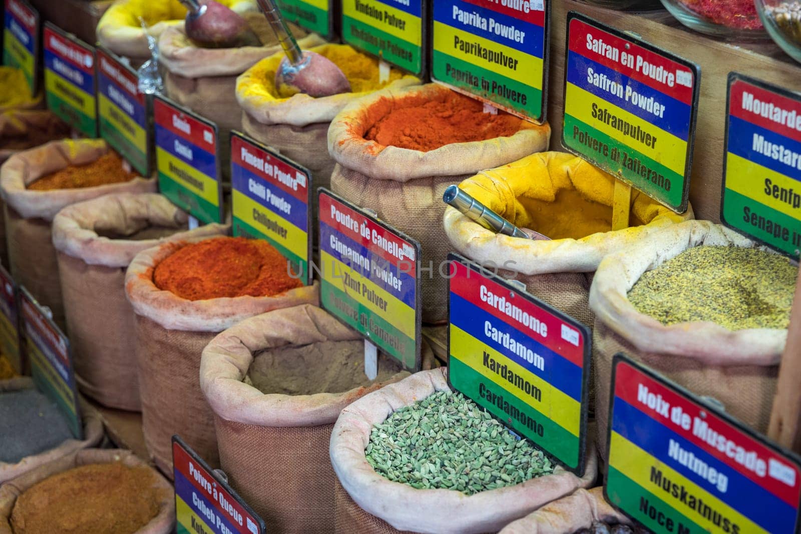 A colorful display of spices and herbs, including cumin, coriander, and ginger. The spices are arranged in small bags and baskets, and there are signs next to each one indicating their names and uses