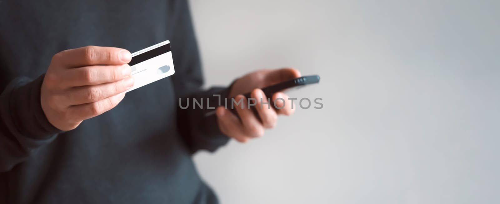 Male hands are holding a mobile phone and a bank card, a young man in a dark casual sweater pays for purchases, makes online banking using an application, close-up view.