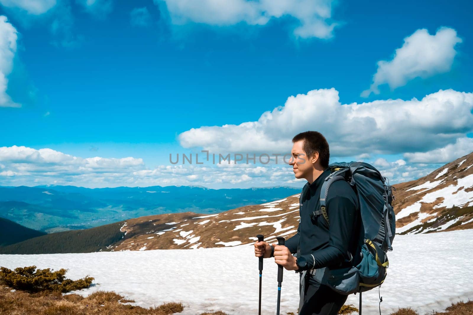 A young man, while hiking in the mountains in winter, looks ahead and admires the stunning mountain landscape. A hiker with tourist equipment climbs to the top of a mountain in a national park.