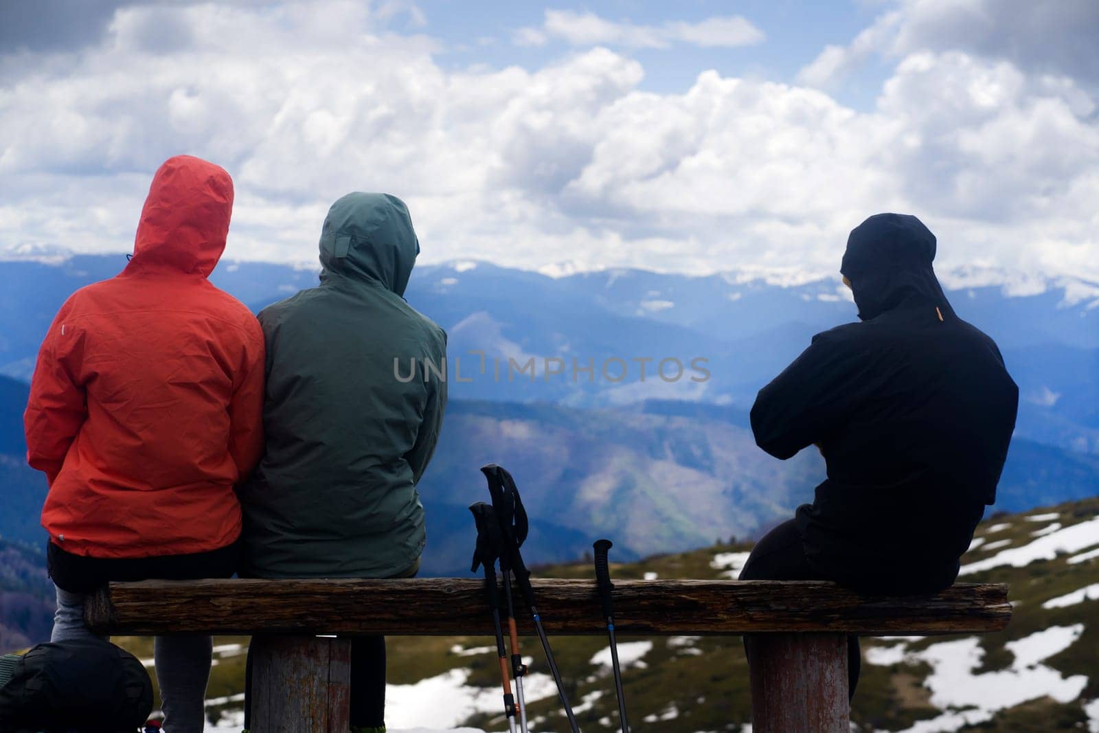 Group of young travelers sit on a wooden bench and enjoy a stunning sunset in snow-capped mountains from a view point. Man and two girls with backpacks and hiking equipment walk in the national park.