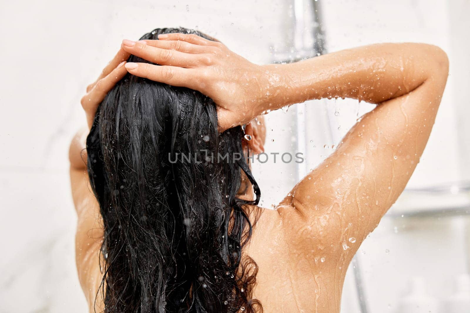 Young woman taking refreshing shower in marble bathroom. Girl washing her hair, back view