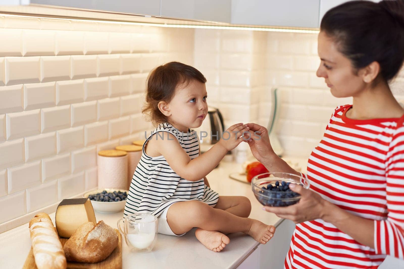 mother gives to the little baby girl fresh blueberries in white kitchen