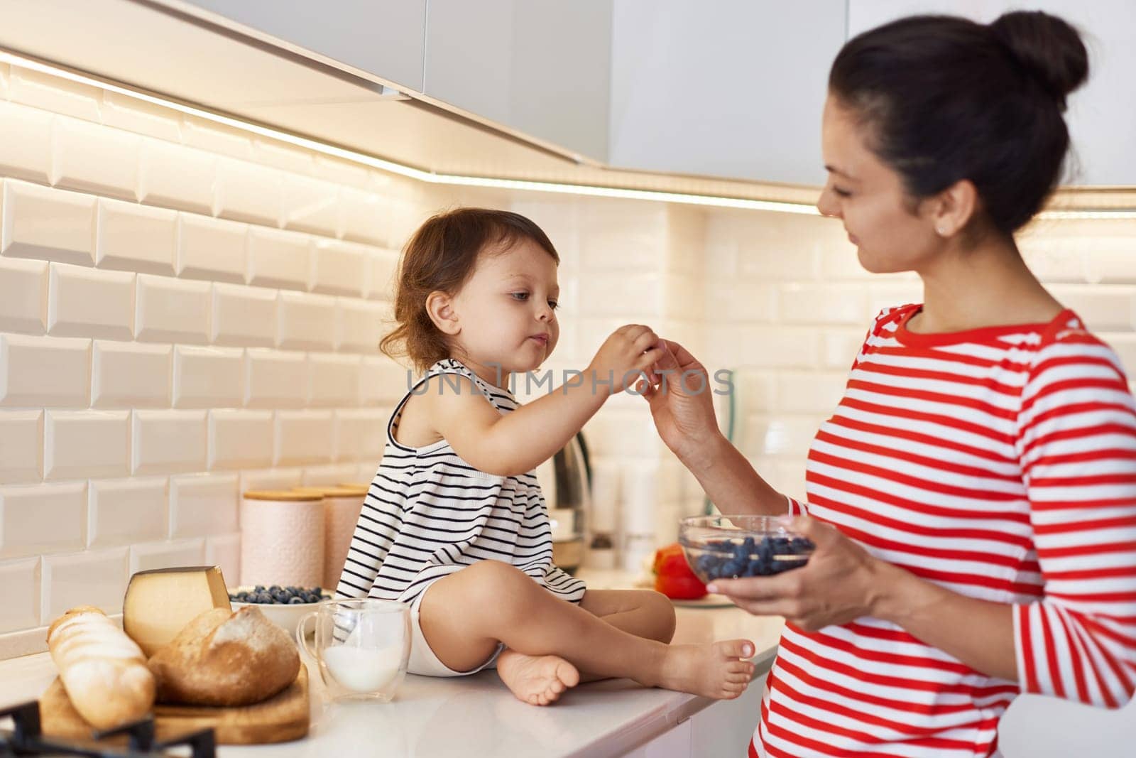 Little baby girl with her mother eating fresh blueberries in white kitchen